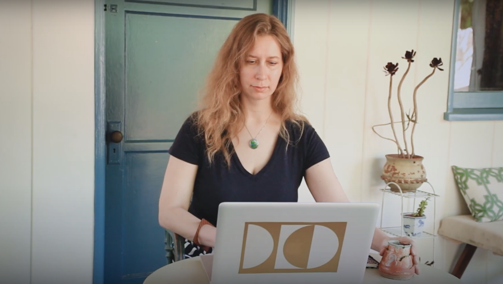 shows author of Neurospicy Housekeeping. She sits at the laptop on a porch, in front of a blue door and white paneling wall with some potted plants. her hair is long and dark, reddish blond, past her shoulder, wavy and lighter toward the bottom. She looks down at the laptop, which has her moon phase logo on it, she wears a navy blue t shirt with low neck and a glass pendant necklace and a brown braclet.